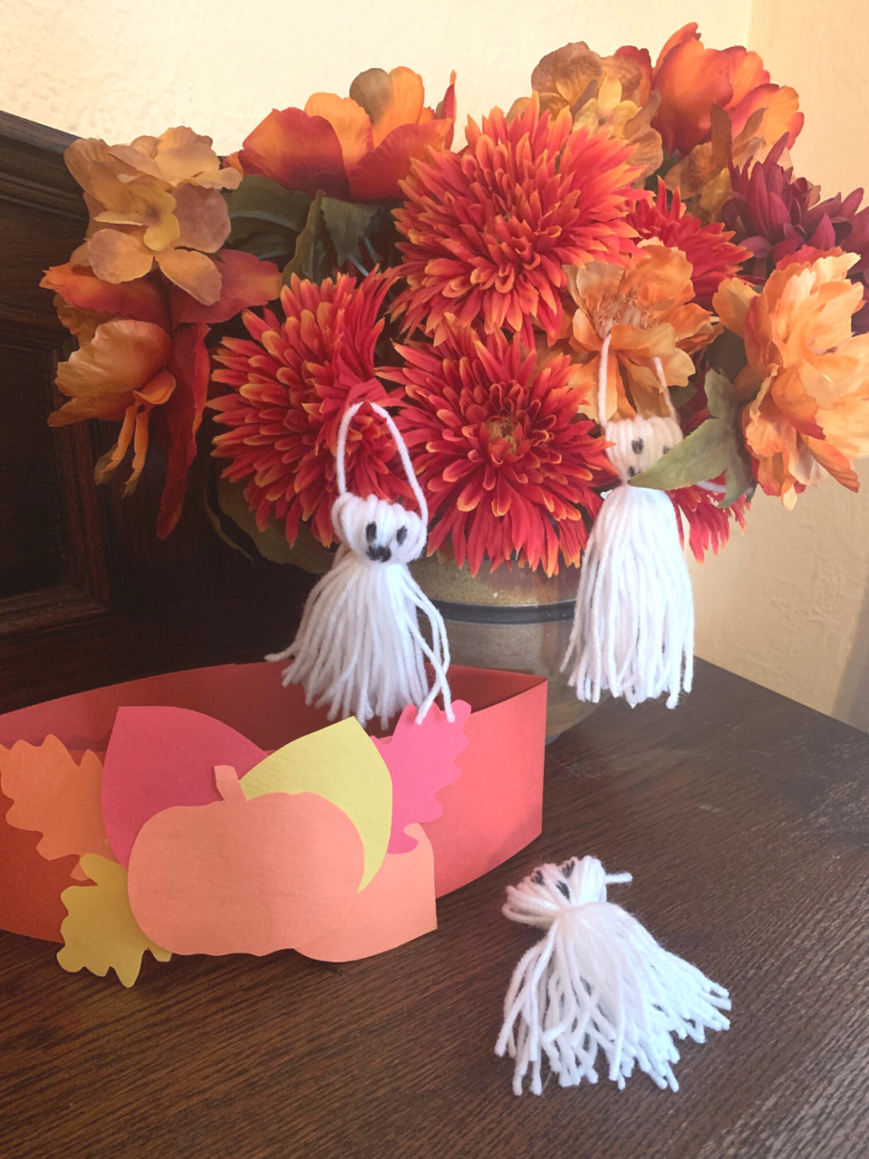 A pumpkin headband and three ghost crafts on a table in front of a fall flower arrangement