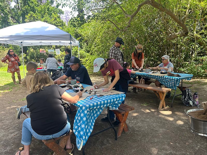 People crafting at outdoor picnic tables under the trees.